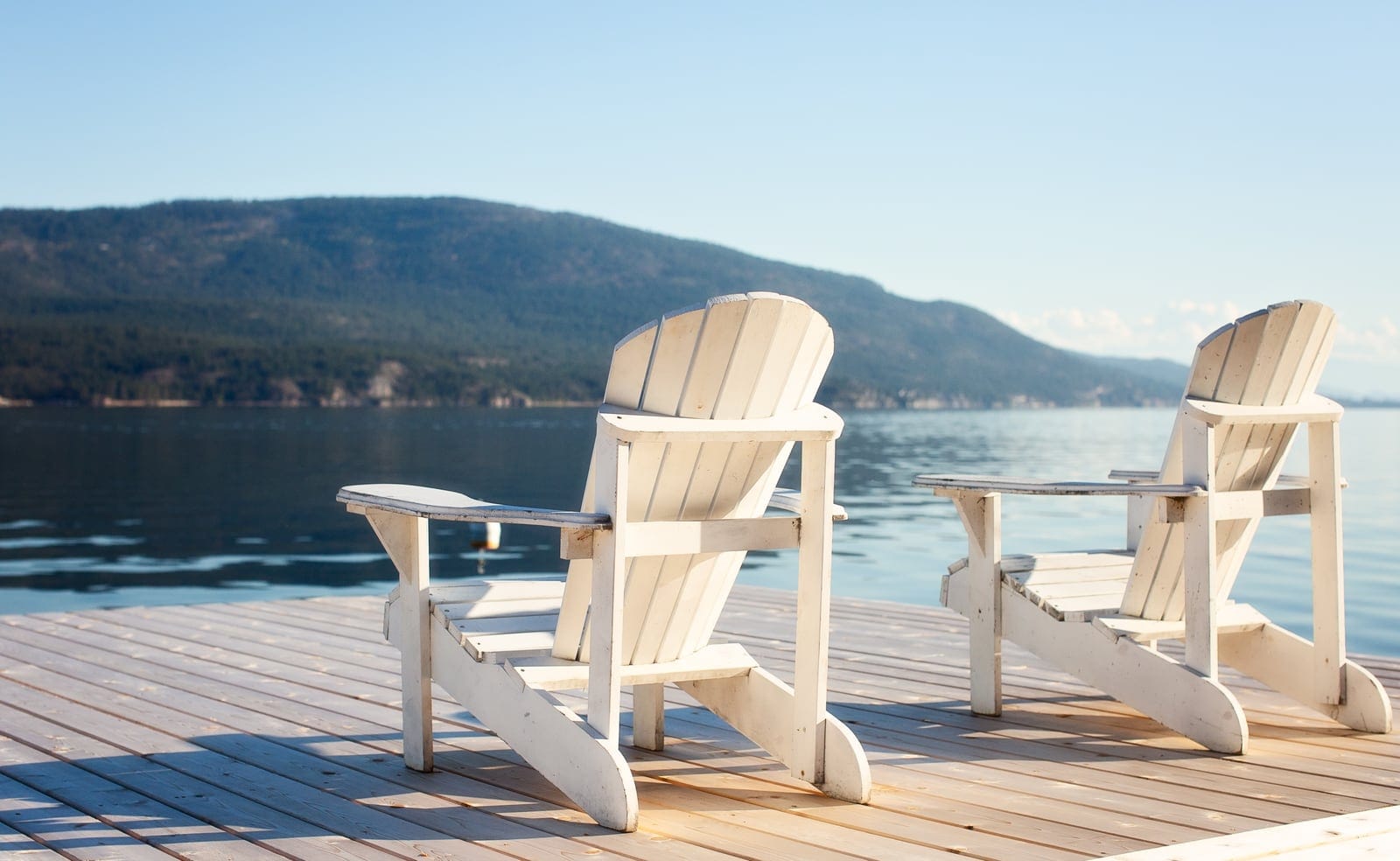 Two white Adirondack chairs on a sunny dock facing a calm lake and forested hills